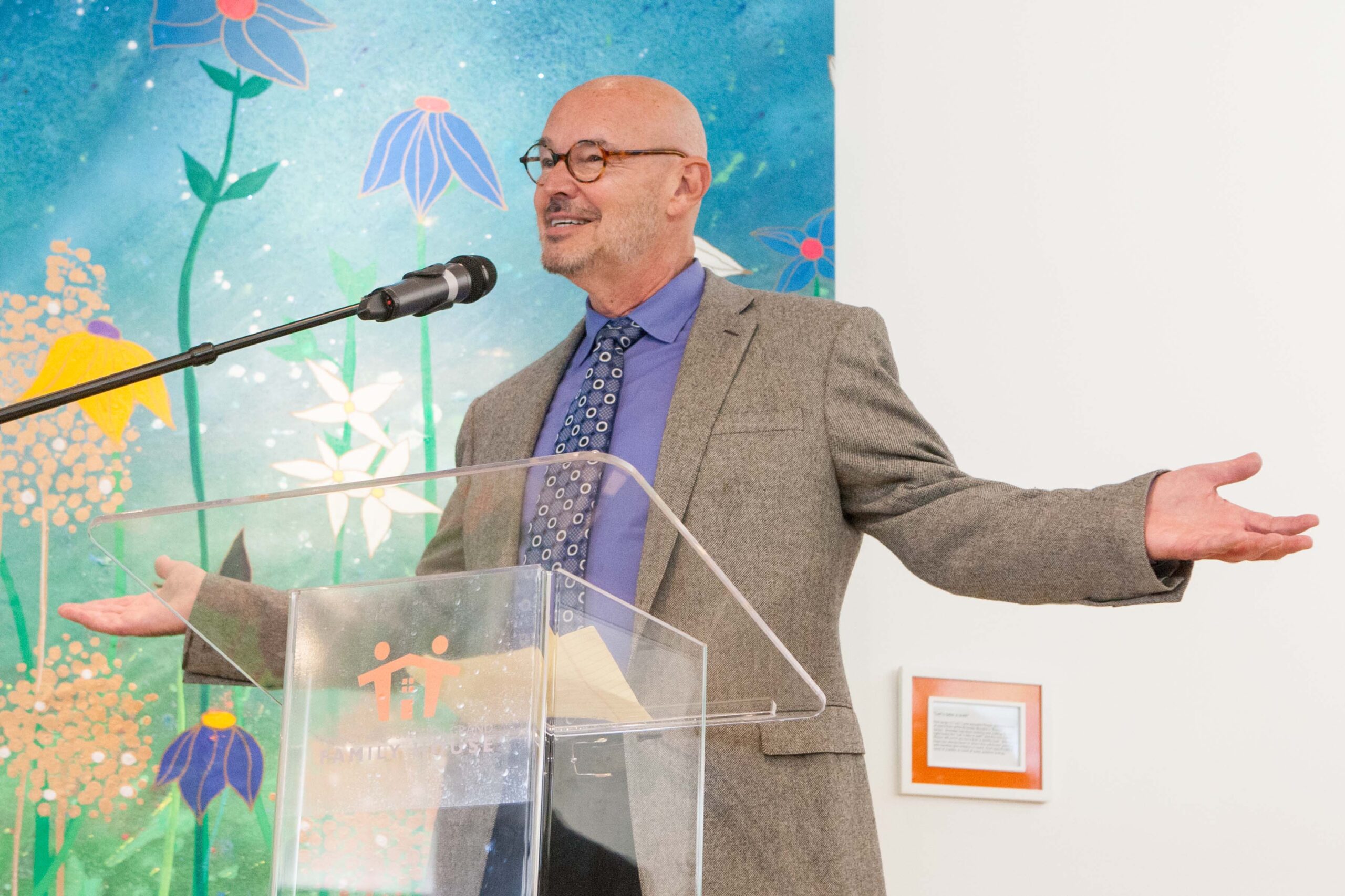 Picture of philanthropist Stephen Grand, bald and wearing glasses in blue tie and collared shirt with grey sportcoat, arms open and speaking into a microphone at the grand opening of the Nancy and Stephen Grand Family House in San Francisco's Mission Bay