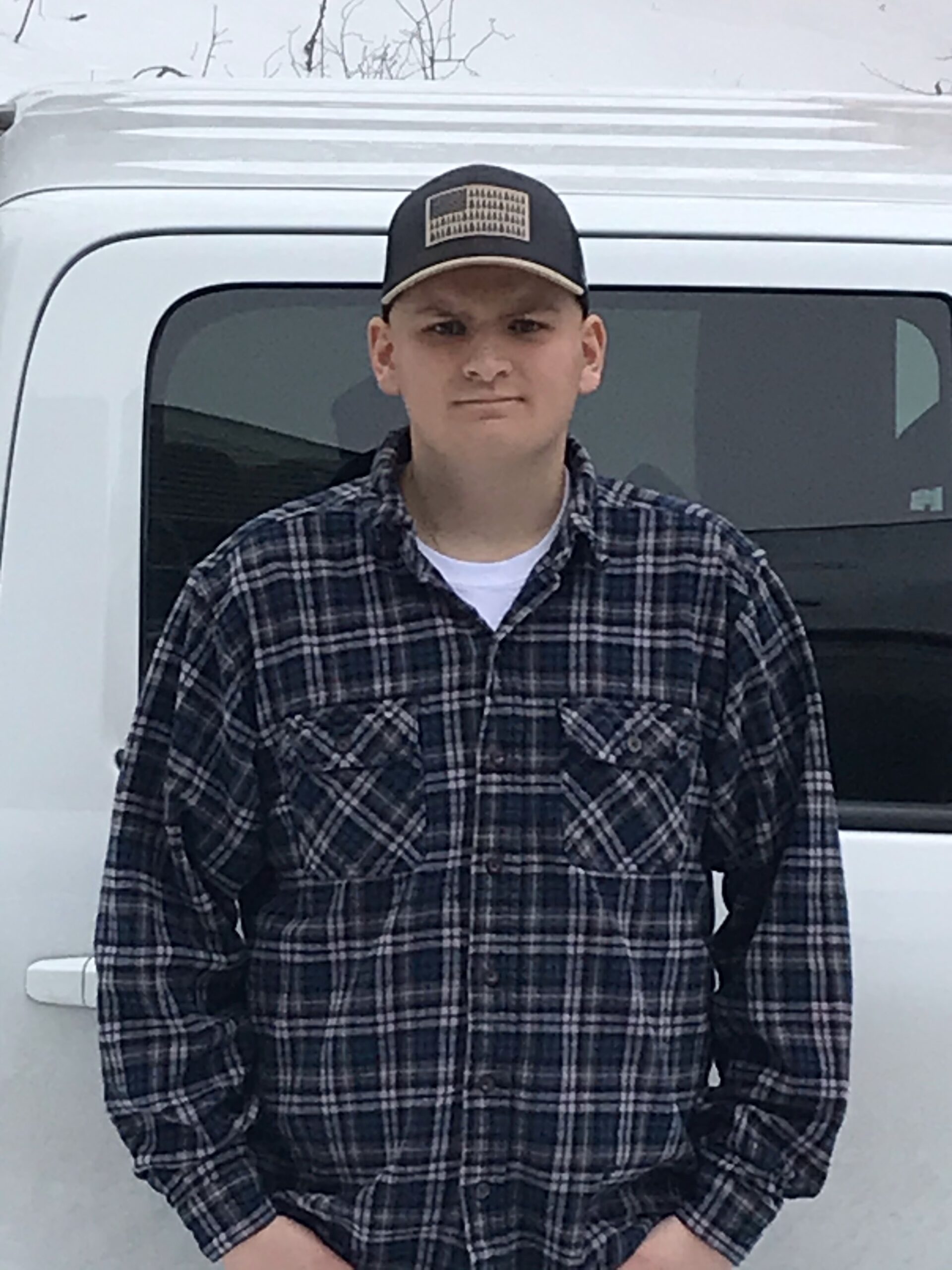Picture of white male named John who is standing with a plaid long sleeve shirt and wearing a ball cap with the american flag on it.